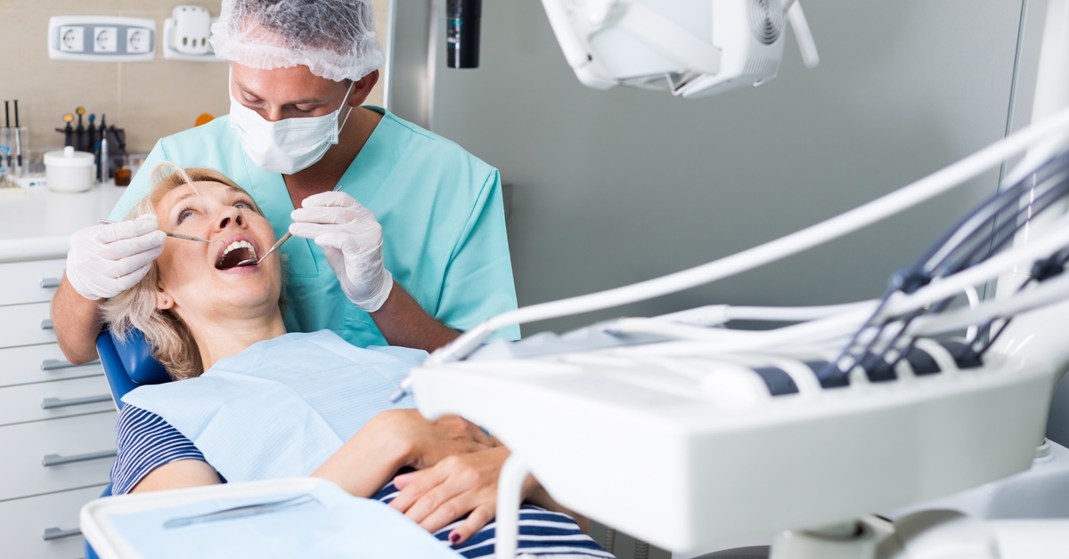 A dentist wearing a mask examines an older woman’s teeth in a dental chair with equipment visible in a modern exam room.