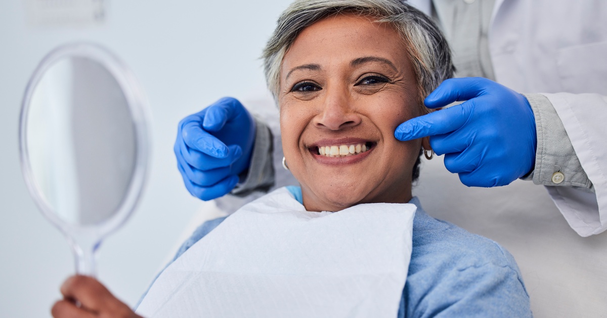 A smiling older woman holds a mirror during a dental visit while a dentist in blue gloves adjusts her cheeks gently.