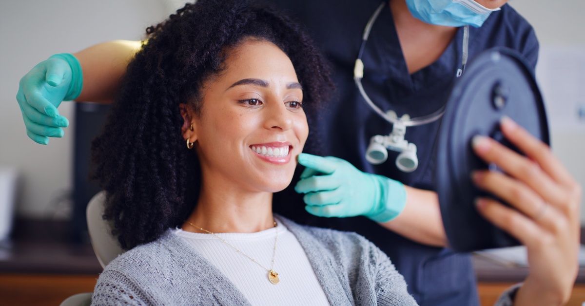 A woman smiling at her reflection after dental treatment while a dentist in gloves points to her mouth.