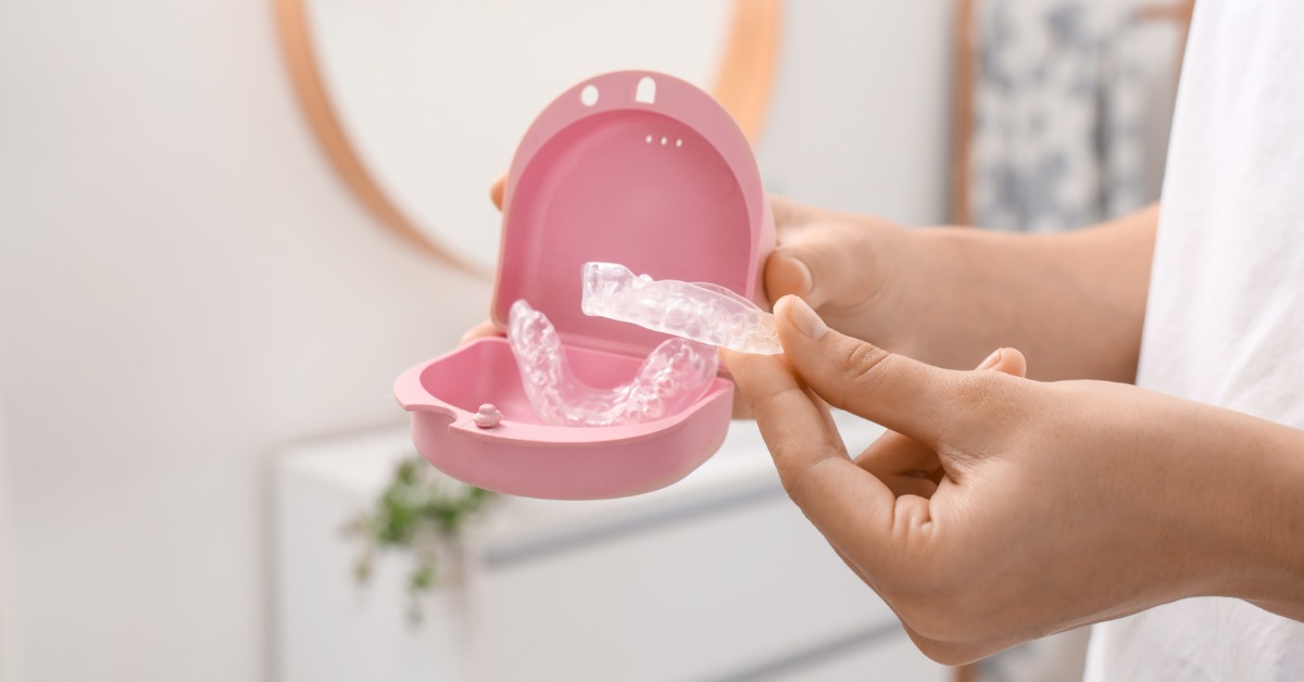 A person holding clear aligners beside an open pink storage case with a round mirror in the background.