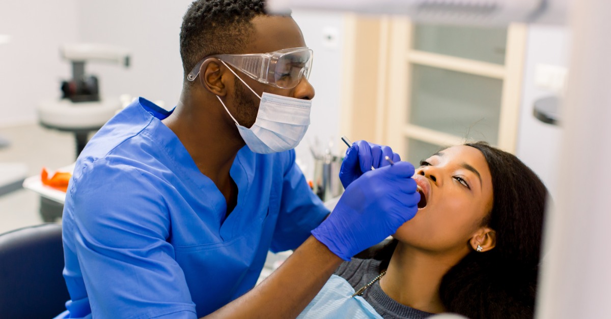 A dentist wearing protective gear examines a female patient’s teeth during a dental checkup in a modern treatment room.