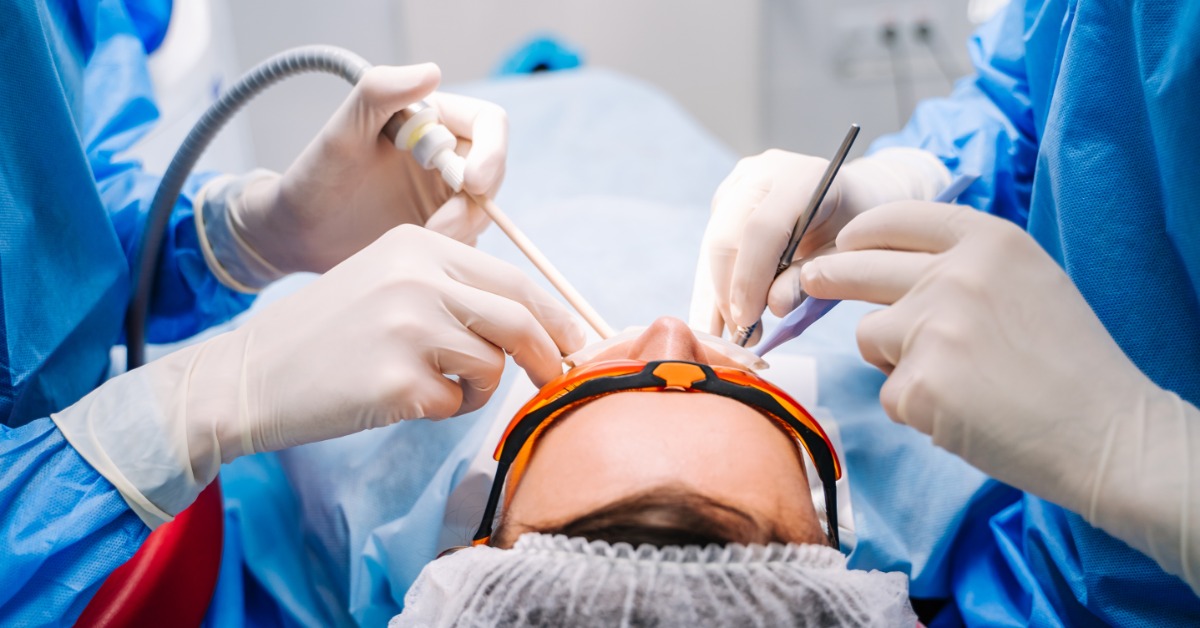 Dentists perform a dental implant procedure on a patient wearing protective glasses while surgical tools surround the mouth.