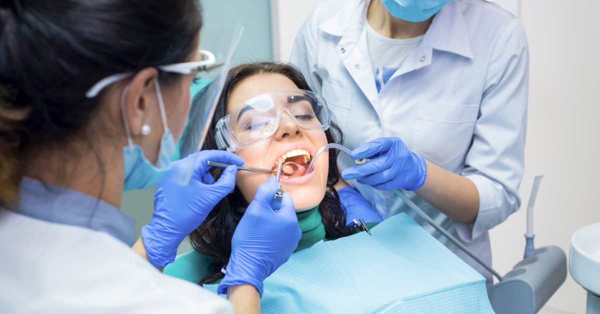 Dentist performs a dental implant procedure while an assistant uses suction as a patient wearing protective glasses reclines.