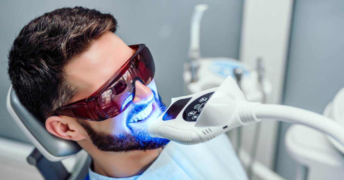 Man wearing protective glasses receives professional teeth whitening with a blue LED light while reclining in a dental chair.