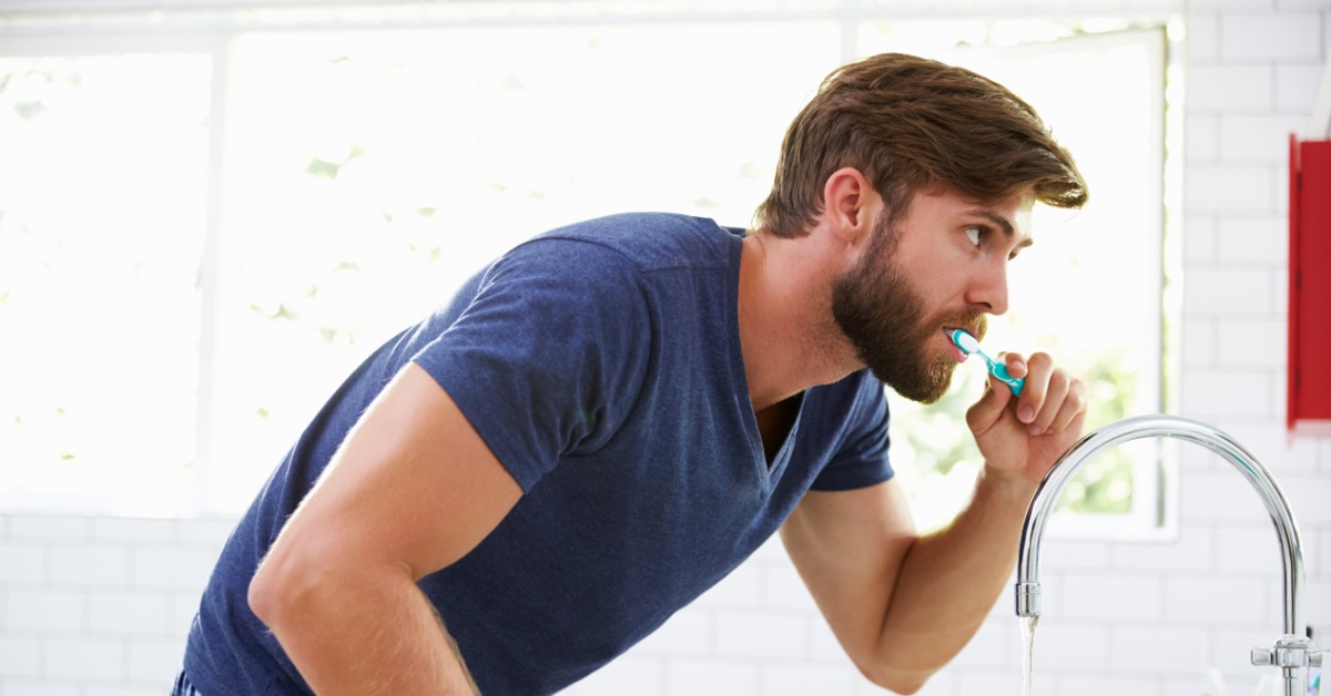 Man brushing his teeth while leaning over a bathroom sink with running water as part of his daily oral hygiene routine.