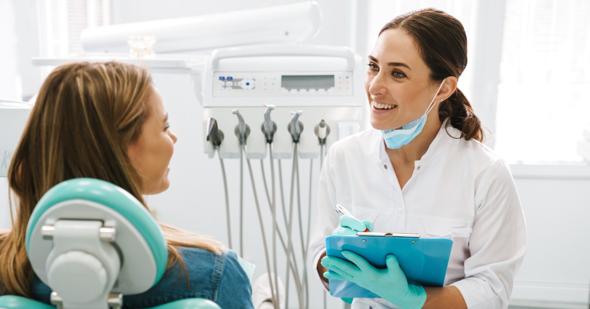Dentist smiling and talking with a patient seated in a dental chair while holding a clipboard in a bright clinic.