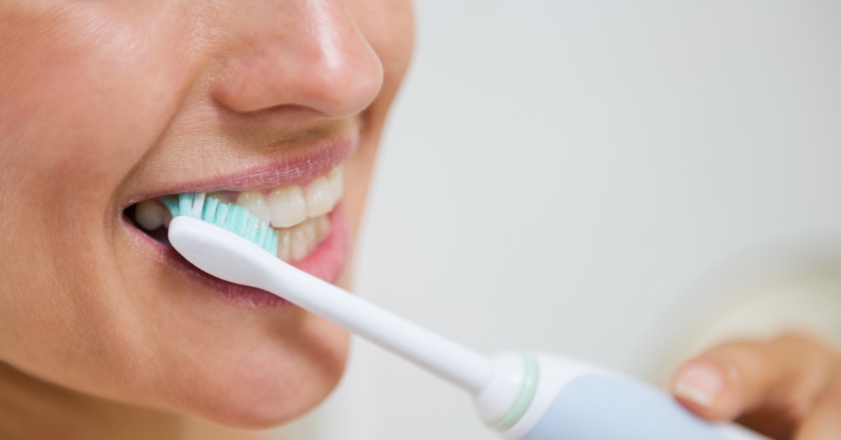 Close-up of a person brushing their front teeth with a blue and white electric toothbrush against a light background.
