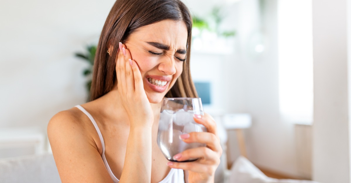Woman winces and holds her cheek while gripping a glass of ice water, showing discomfort from cold tooth sensitivity.