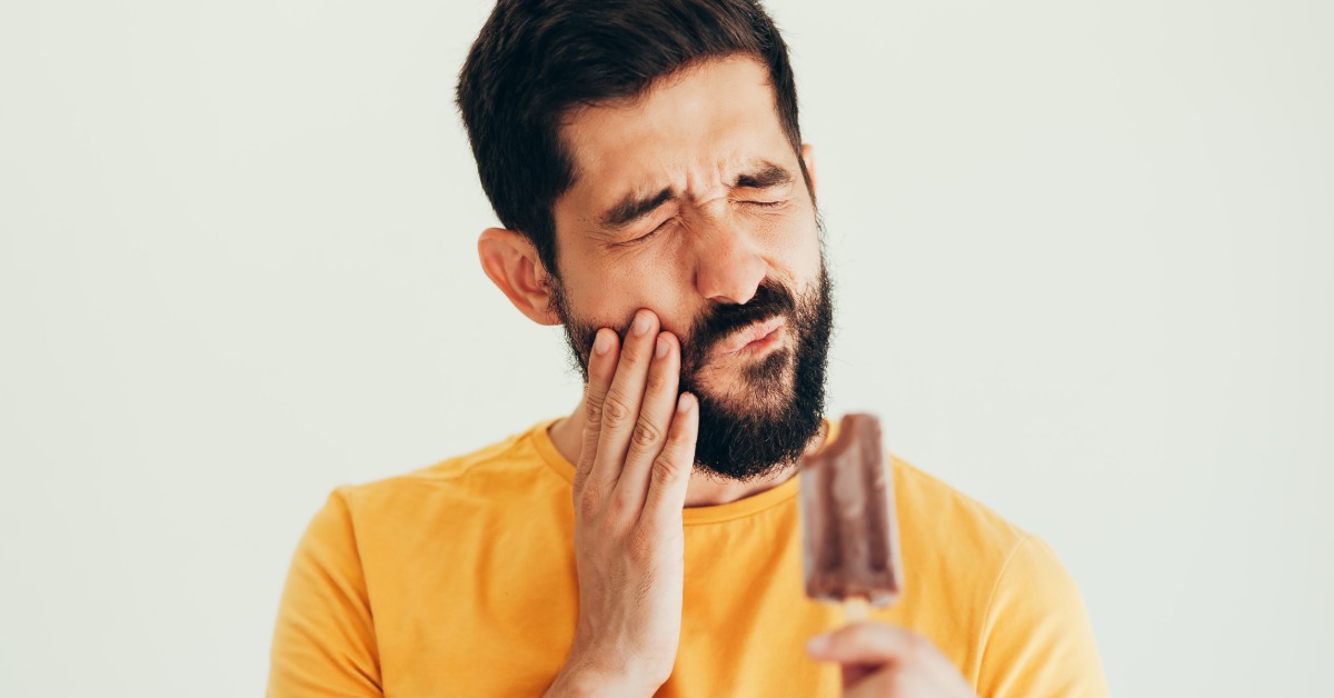 Man winces and holds his cheek while looking at a chocolate ice pop, showing discomfort from cold tooth sensitivity.