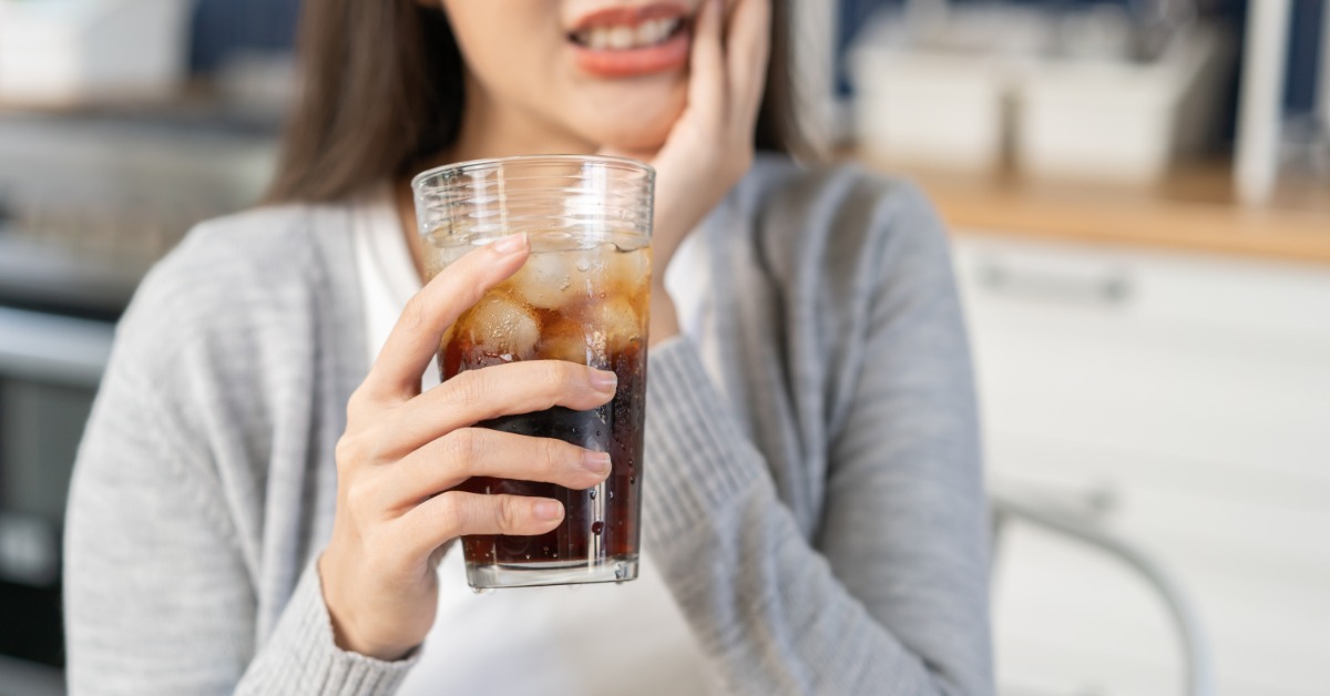 Woman holds her cheek in pain while gripping a glass of iced soda, showing discomfort from cold tooth sensitivity.