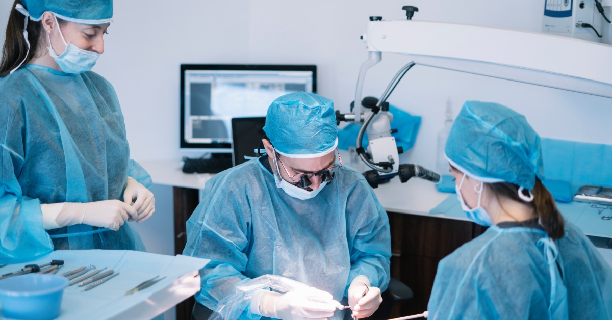 Dental team in surgical gowns and masks performs a dental procedure on a patient in a clinical treatment room.