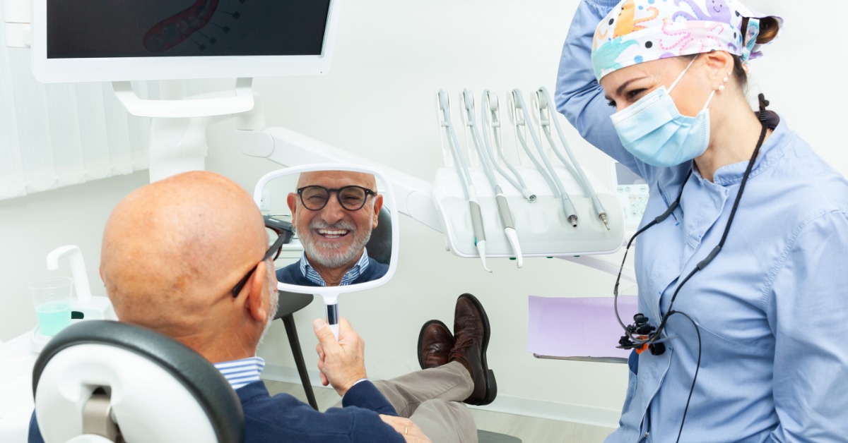Smiling older dental patient looks at his teeth in a hand mirror while a masked dentist sits beside him.